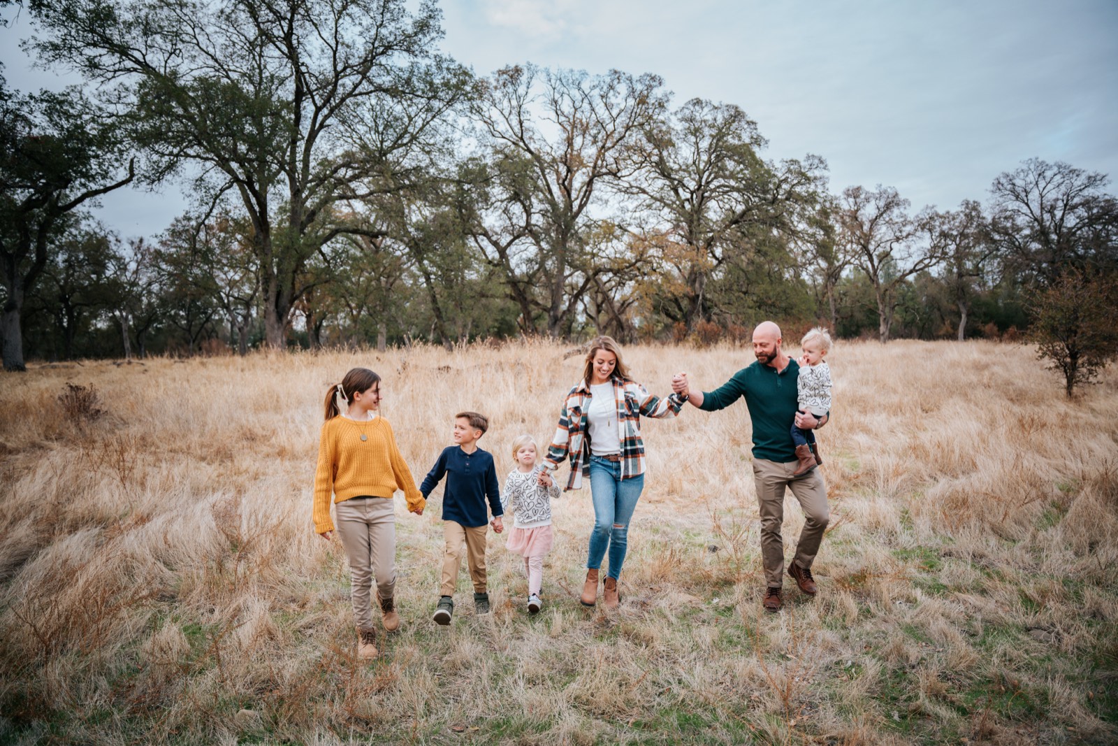 Ashley and her family walking hand in hand through a field of golden grass under oak trees