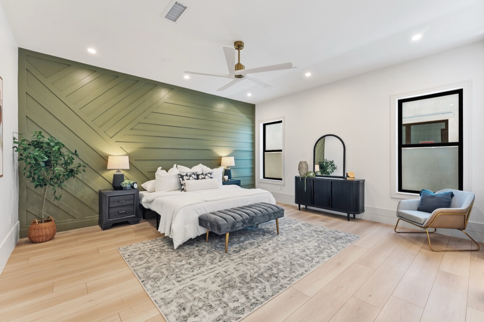 A staged bedroom with a green paneled accent wall, light wood floors, and black-framed windows